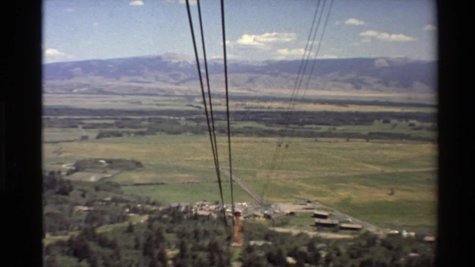 1980s 8mm Western U.S. cable car view looking over pasture, trees, small structures, and distant mountain ranges.