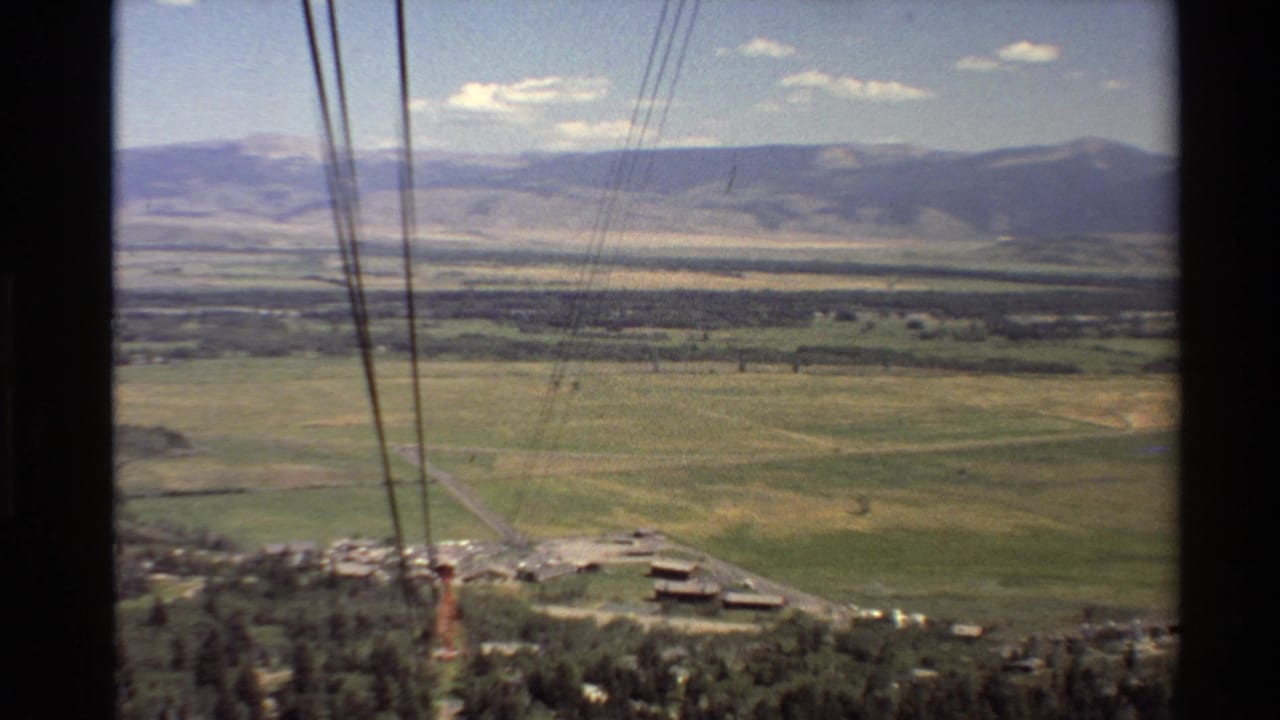 Cable car ride footage shows foreground cables crossing a Western U.S. valley of fields, trees, roads, and mountains.