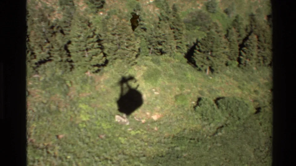1980 Western USA 8mm frame showing a helicopter shadow over a grassy mountain slope with evergreen trees above.