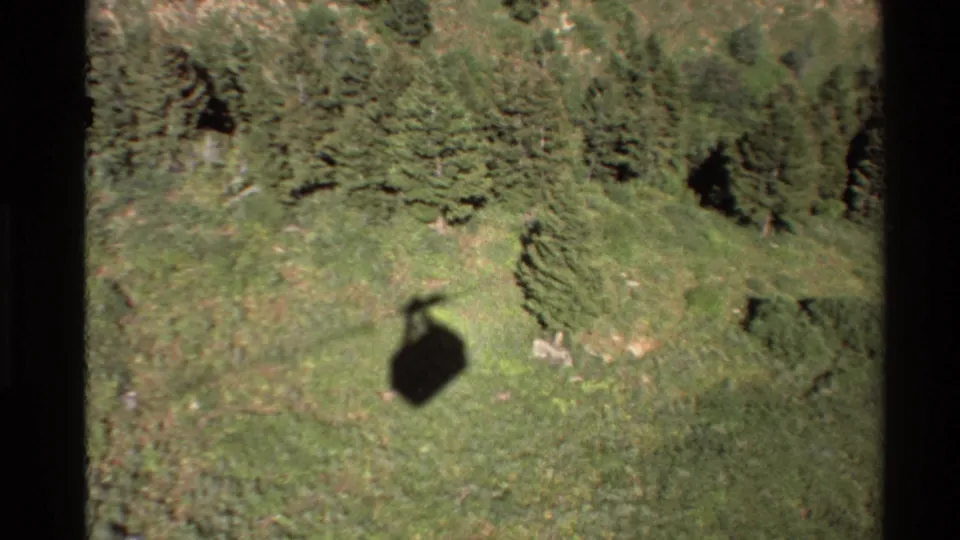 Vintage 8mm aerial frame with a helicopter shadow crossing a grassy mountain area near evergreen trees.