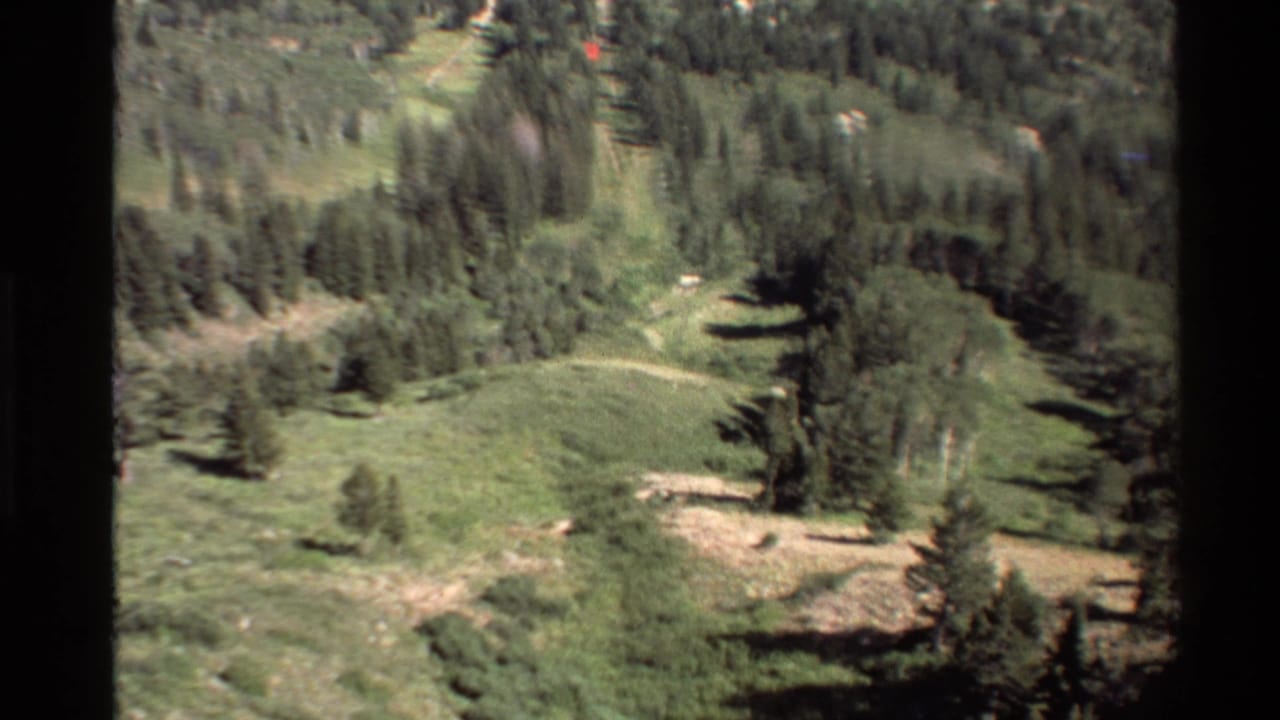 1980s 8mm home-movie view over green mountain slopes, scattered trees, and open meadow in the Western USA.