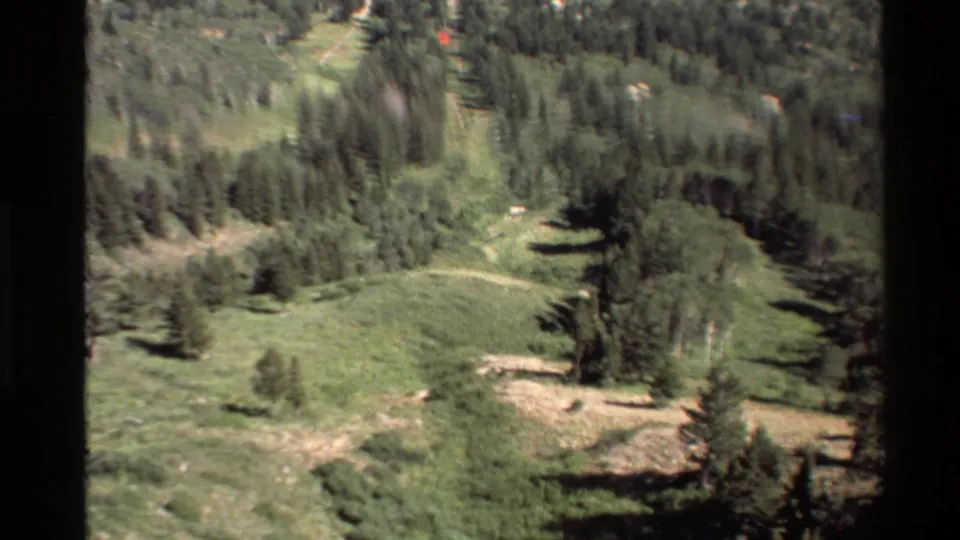 1980s 8mm home-movie view over green mountain slopes, scattered trees, and open meadow in the Western USA.