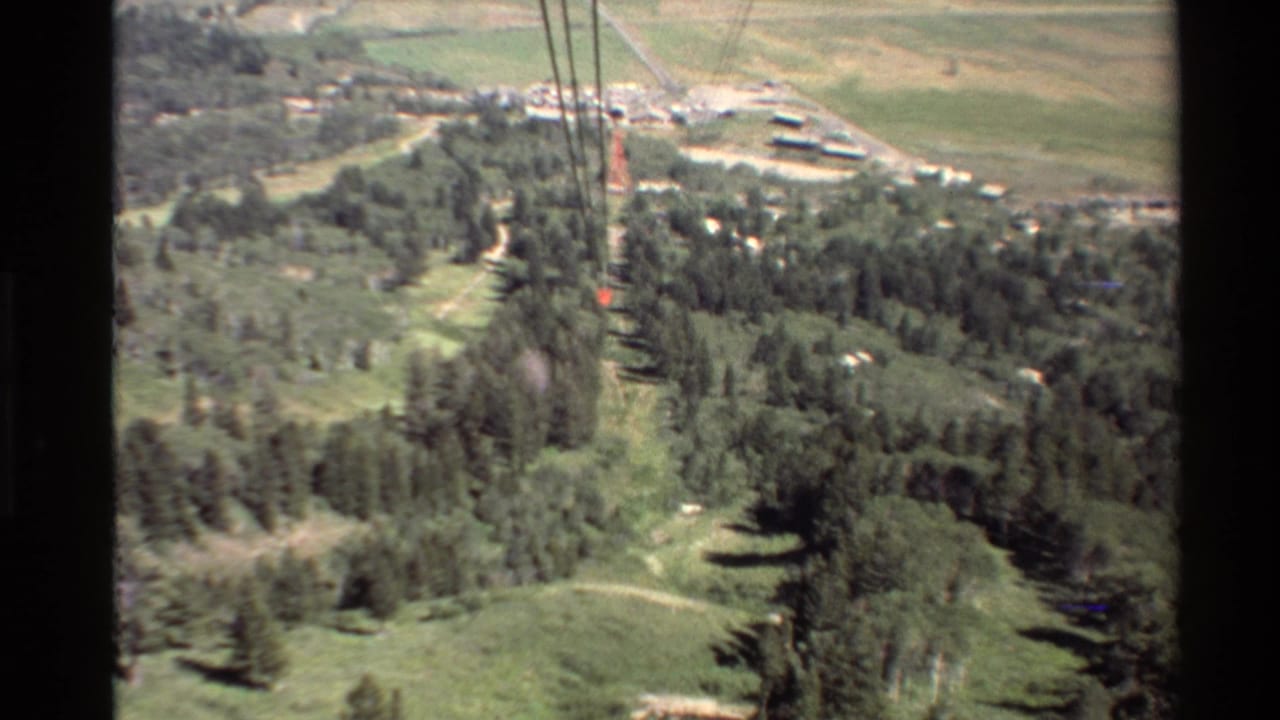 Cable lines pass over a green valley with trees, meadows, roads, and distant buildings.