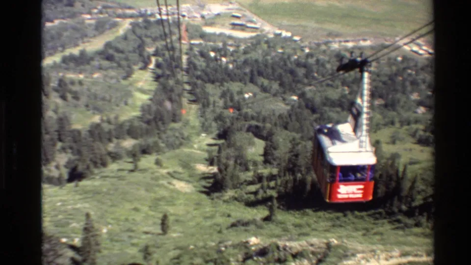 Red aerial cable car traveling down suspended lines over a green mountain landscape in 1980s Western USA home-movie footage.