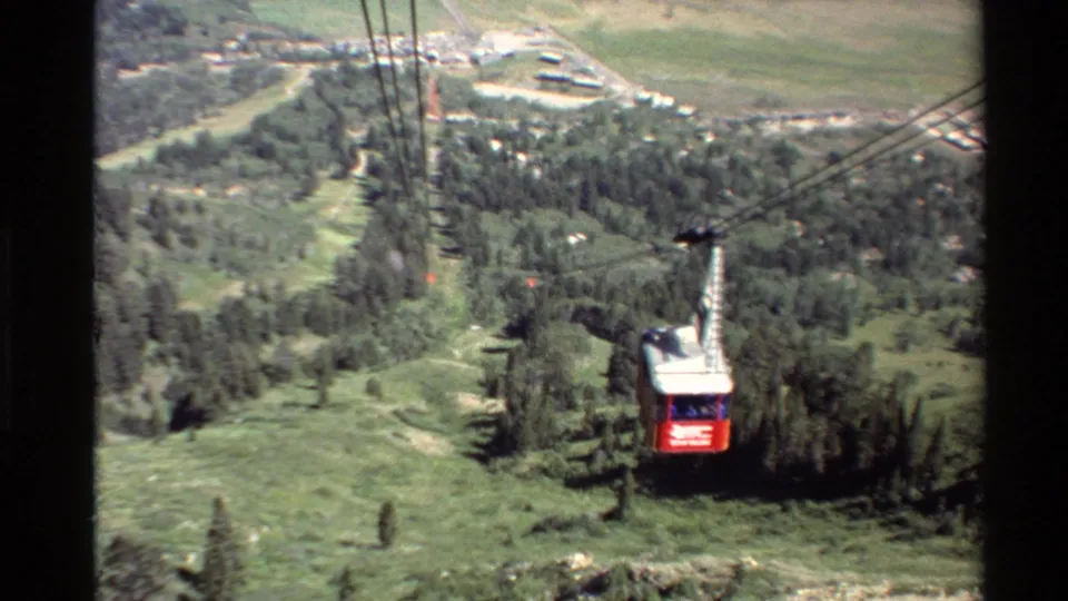 Red cable car seen from above or behind as it moves over trees, meadow, and valley terrain in Western USA 8mm footage.