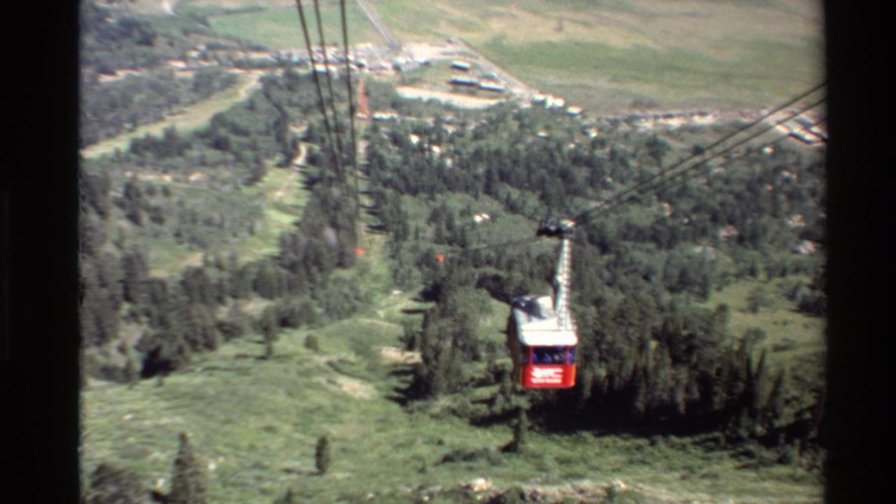 1980 Western USA 8mm frame showing a red and white aerial tram car suspended over evergreen slopes and a valley below.