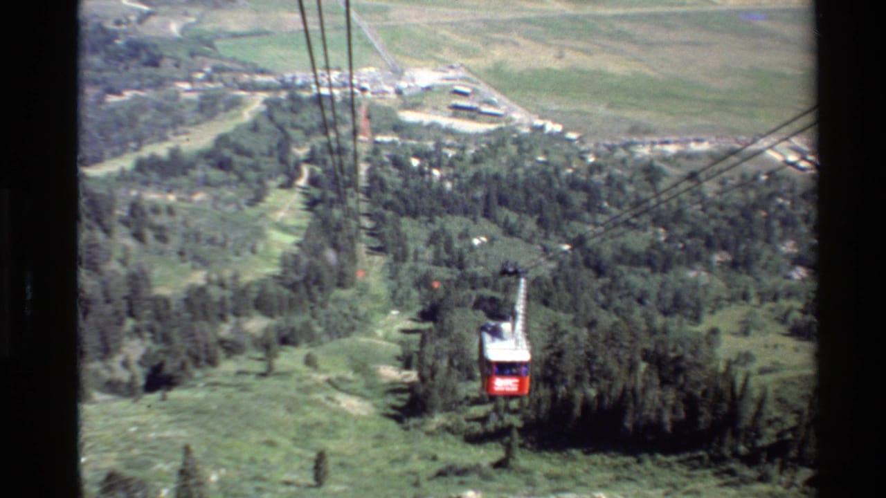 The tram car continues along the cable line above wooded slopes, with pale roads and a valley settlement visible beyond.