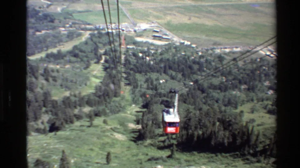 The tram car continues along the cable line above wooded slopes, with pale roads and a valley settlement visible beyond.