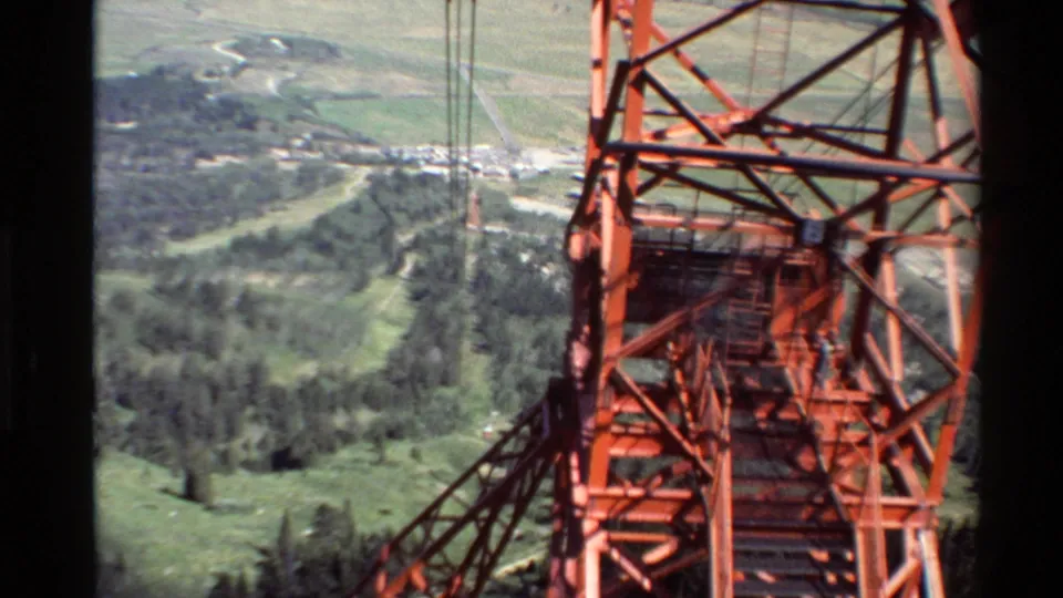 Close view of the orange tram support tower and cable structure, with forested slopes and valley roads in the background.