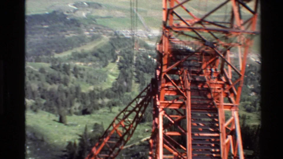 1980 Western U.S. 8mm view of a red cable support tower passing close to the camera above green forested hills and distant plains.