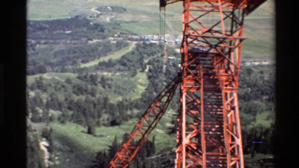 A close aerial frame shows the red lattice tower and diagonal support arm with forested slopes and open land beyond in 1980 8mm footage.