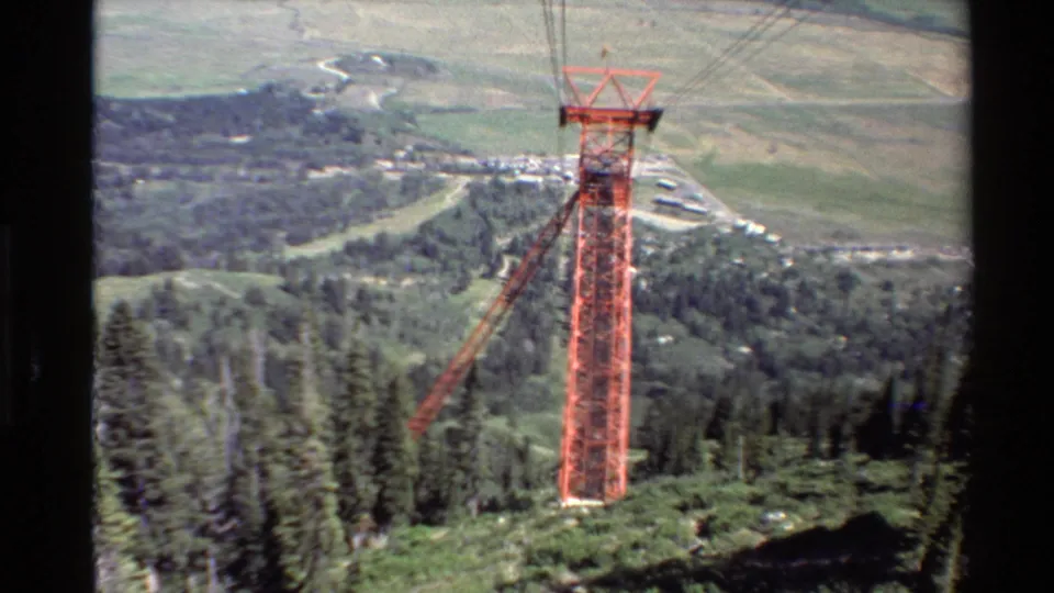 The camera has pulled back to show more of the red cable tower standing above evergreens, with hills, plains, and a small settlement in the background.