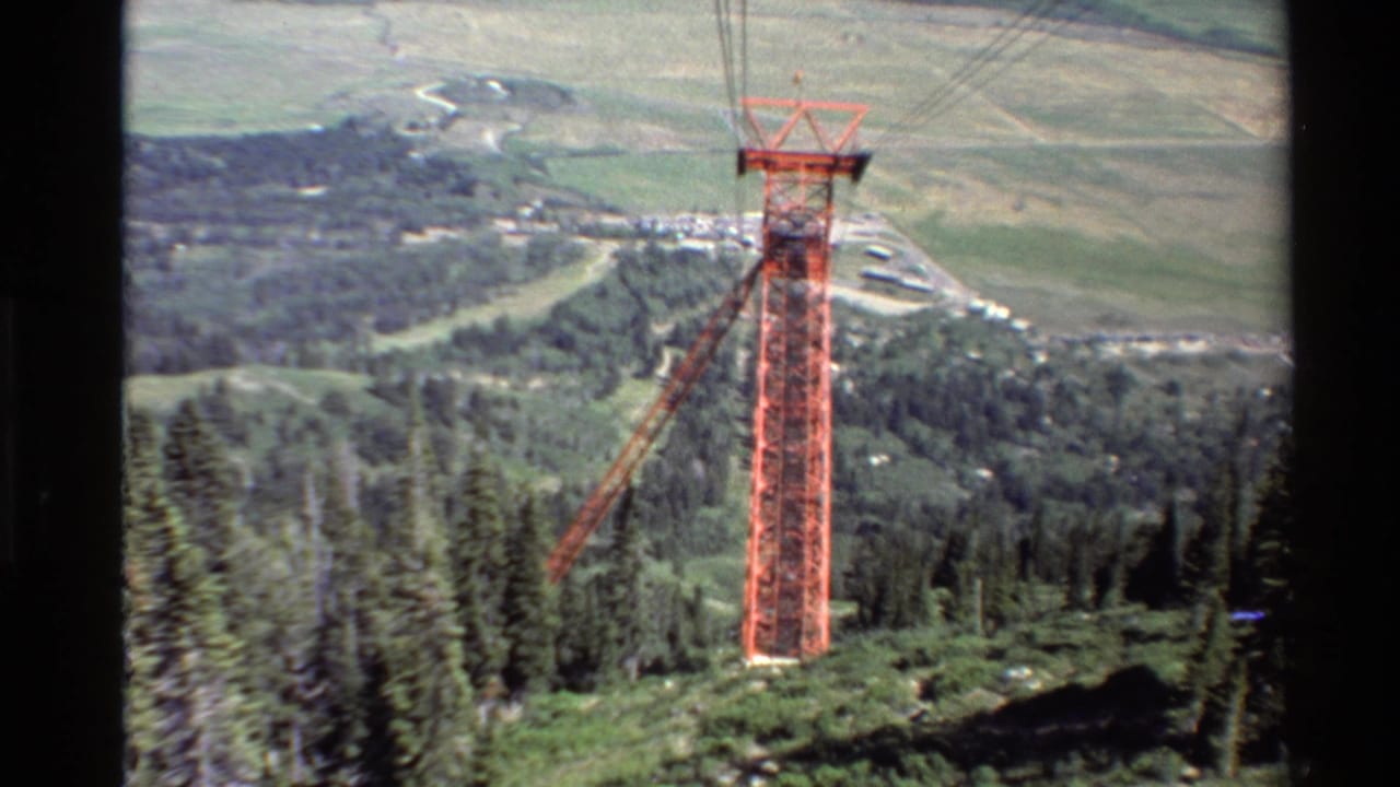 1980s 8mm view from a chairlift approaching a red ski-lift tower over pine trees and a broad Western U.S. valley.
