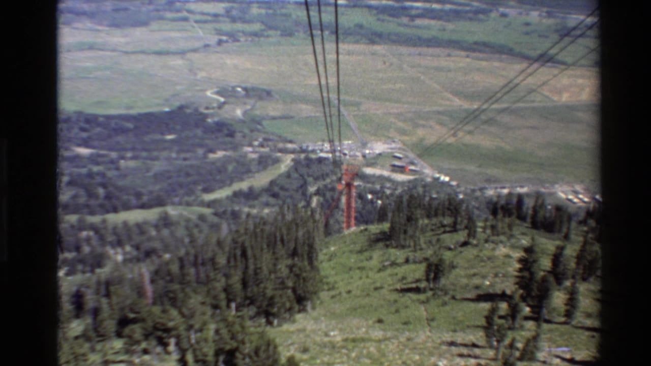 Lift cables stretch over a grassy ridge with trees and a valley below.