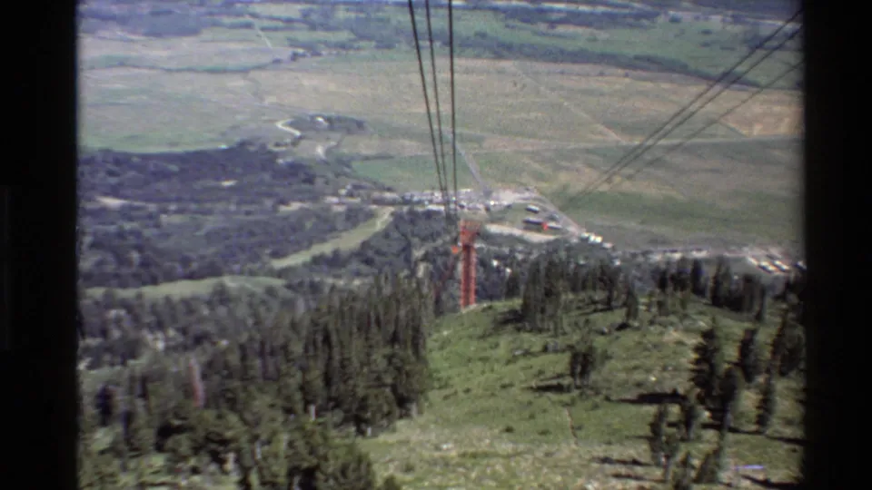 View from the ascending chairlift over a green ski slope, cables, pine trees, and distant valley landscape in the Western U.S.