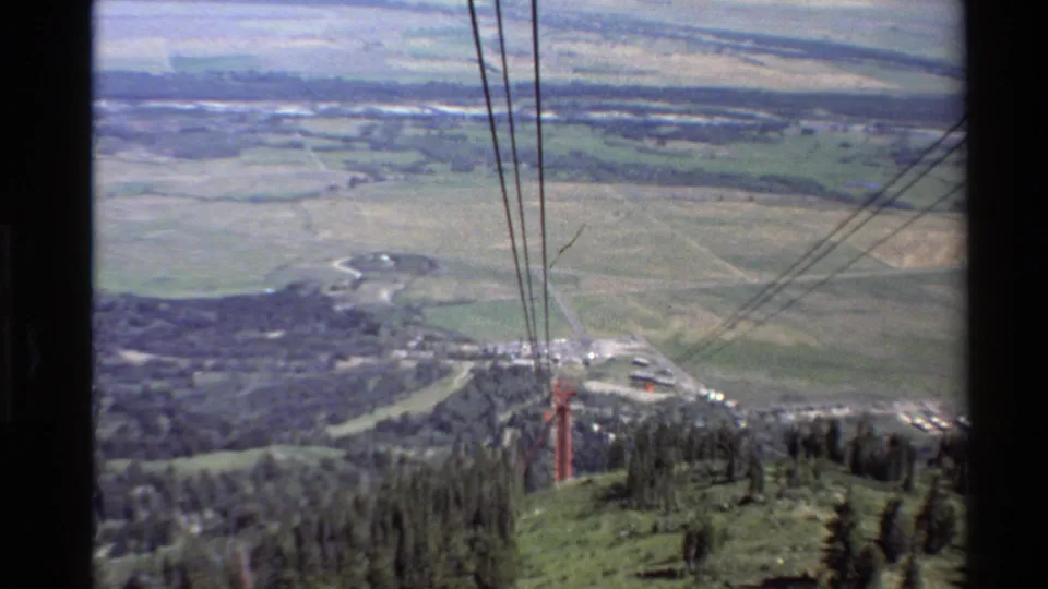 Aerial 8mm chairlift perspective over summer ski terrain, with cables, forest, roads, and fields visible below.