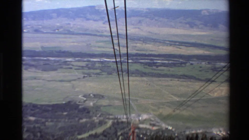 Final chairlift view looking outward over cables, muted green slopes, roads, and a distant mountain valley landscape.