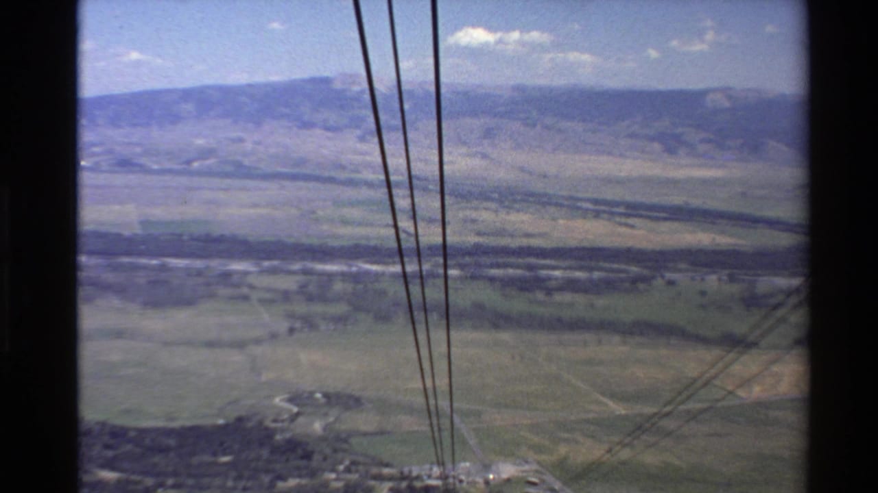 1980s 8mm Western U.S. footage showing cable lines over green valley fields and distant mountains from a high tramway viewpoint.