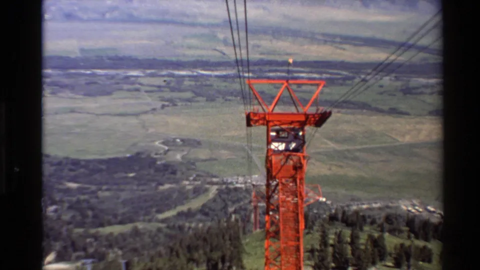 1980s 8mm view of a red tramway support tower framed against fields, forested hillside, and distant mountain terrain.