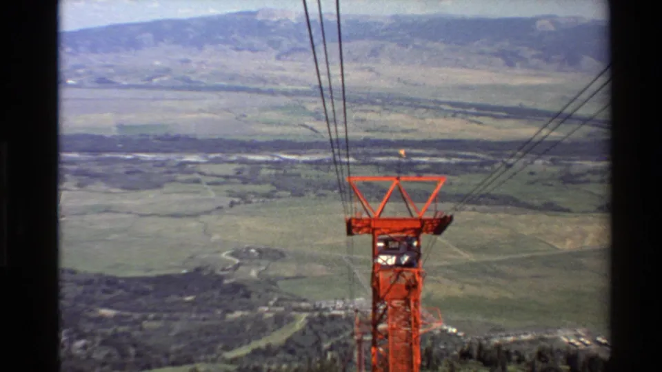 High cable car viewpoint over a Western U.S. valley, with a red support tower, black cables, fields, and mountain ridgelines.