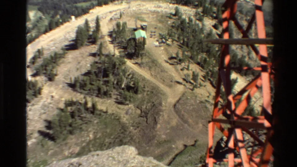 High view of rugged Western USA terrain with a red lattice tower, dirt roads, scattered trees, and a small green-roofed building.