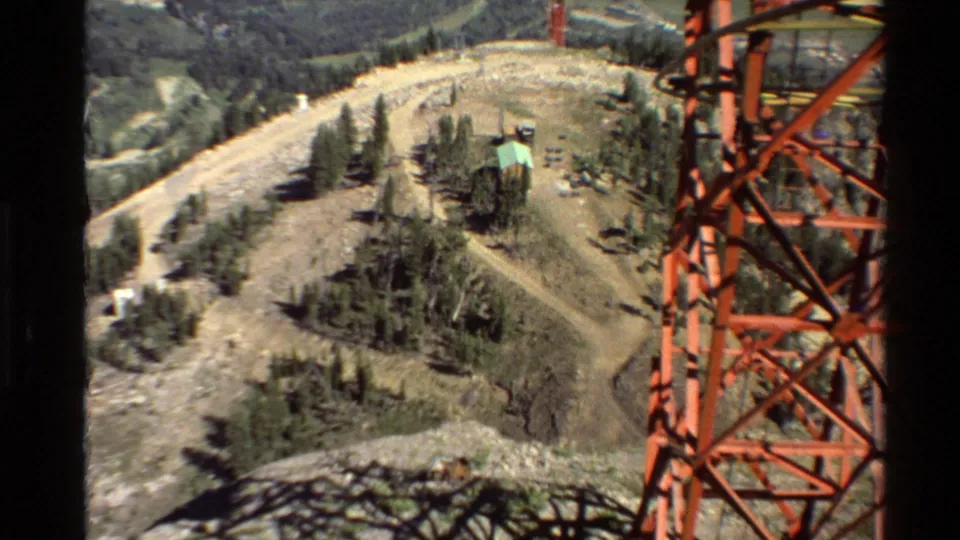 8mm view across a rocky slope in the Western USA, with trees, a dirt road, a small structure, and red tower steel at the edge.