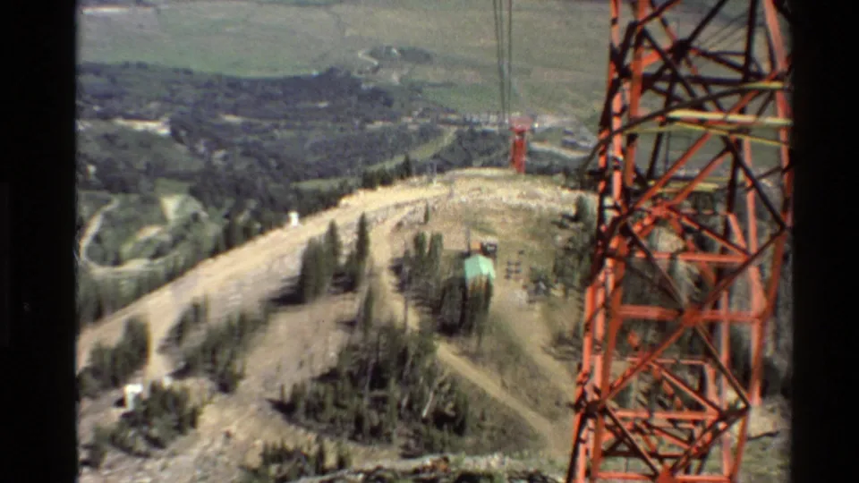 Western USA mountain view with red tower sections, overhead wires, forested slopes, dirt tracks, and a distant valley.