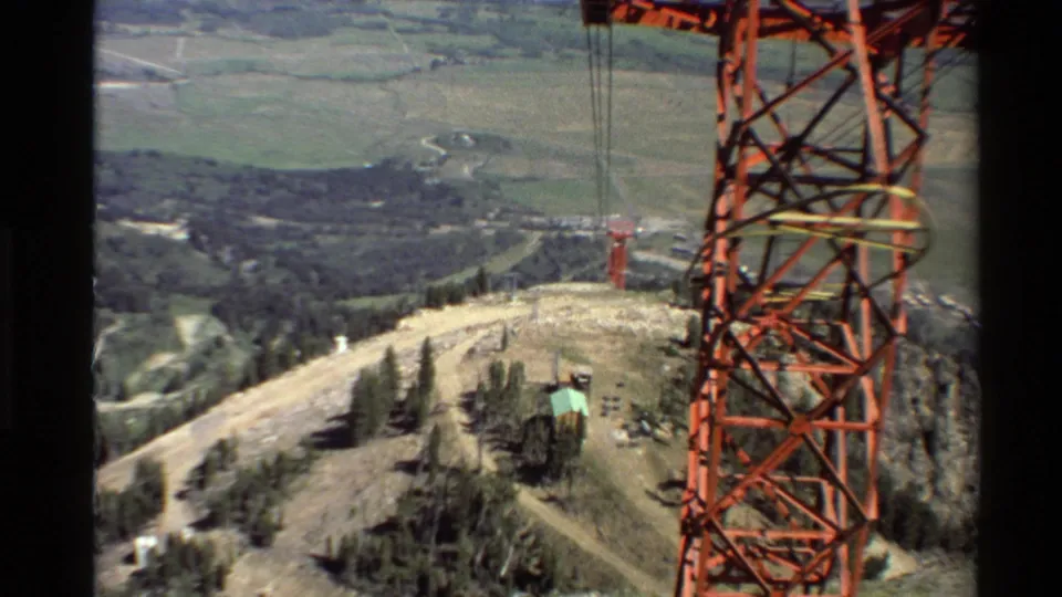 1980s 8mm view of a red electrical transmission tower on a Western U.S. mountain ridge with valleys and evergreens below.