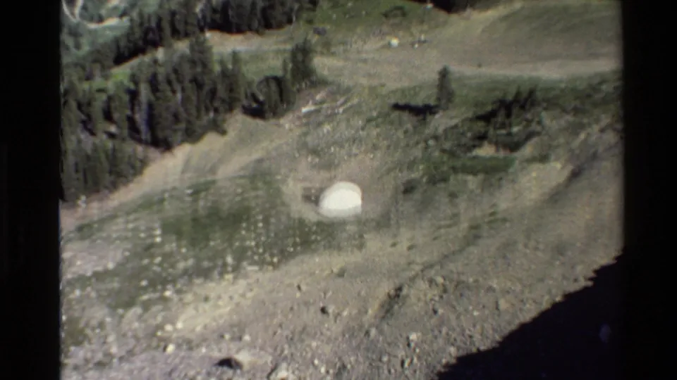 1980s Western U.S. mountain view with evergreen trees, dirt tracks, a pale utility-like structure, and open hillside.