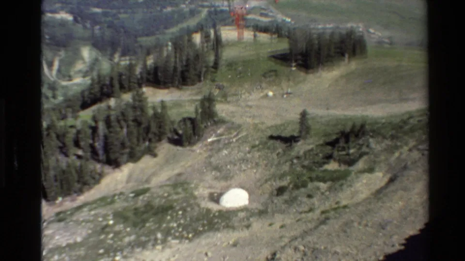 1980 Western USA 8mm view of a rocky hillside below cable lines, with evergreen trees and a small white dome-like structure.