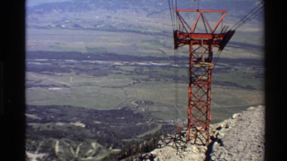 1980 Western USA 8mm frame of a red cable tower on a rocky ridge, with suspended cables and patchwork valley fields beyond.