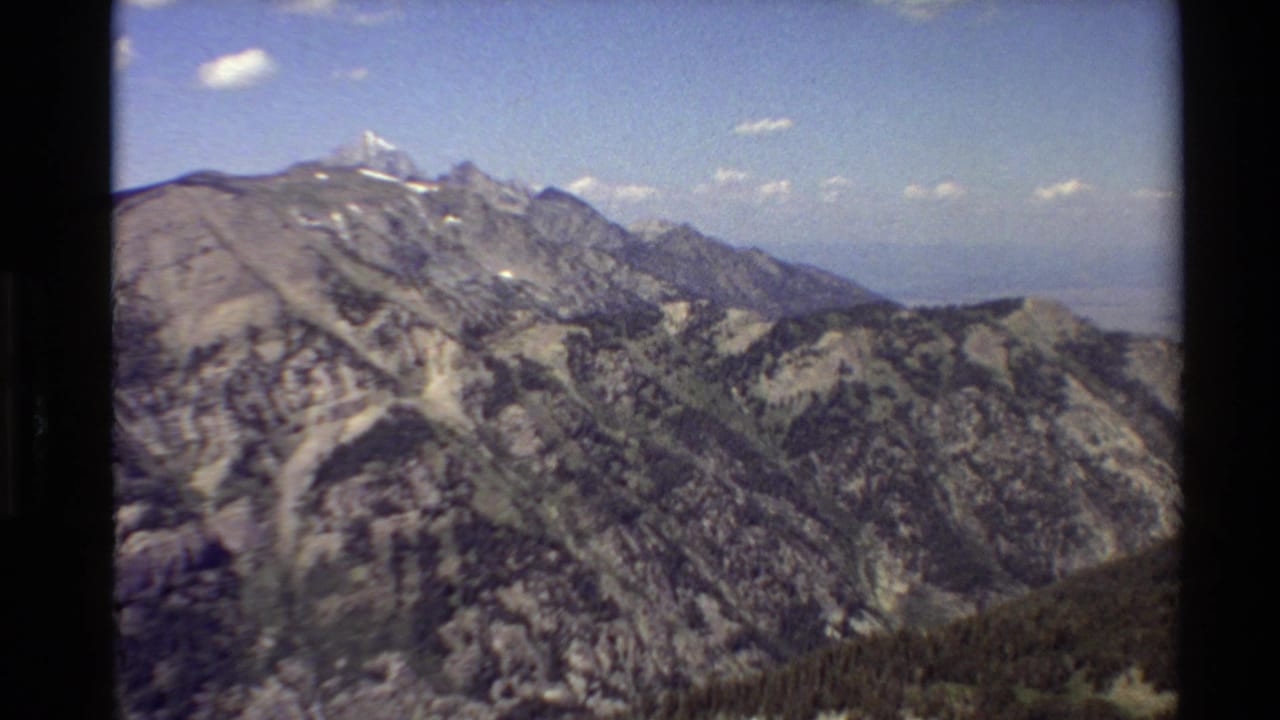 1980 Western USA 8mm frame showing rugged ridges, scattered evergreens, distant snowy peaks, and small clouds in a bright sky.