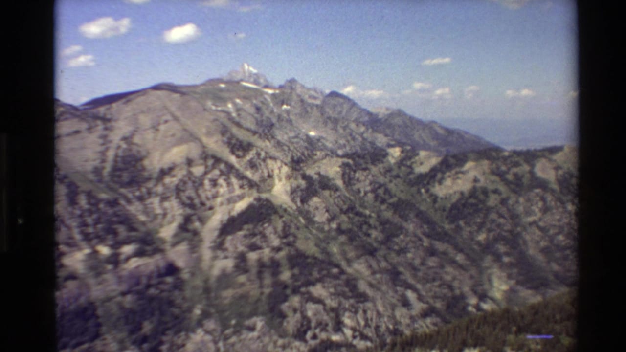 Mountain ridges and forested slopes beneath a blue sky with small clouds and dark film edges.