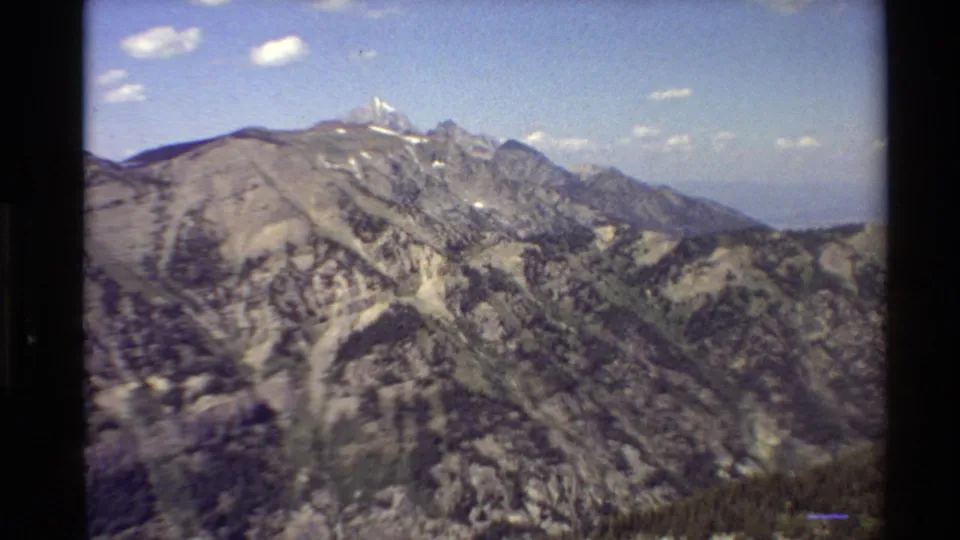 1980 Western USA home-movie frame with rugged mountain terrain, snow on high peaks, wooded slopes, and analog film borders.
