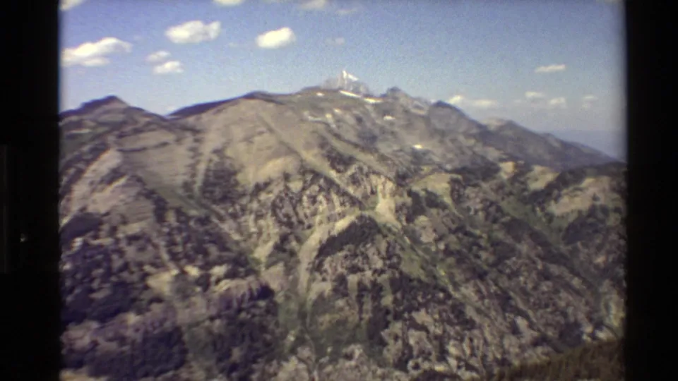 1980 Western USA 8mm frame showing a broad rocky slope leading toward snow-tipped summits and distant mountain layers.