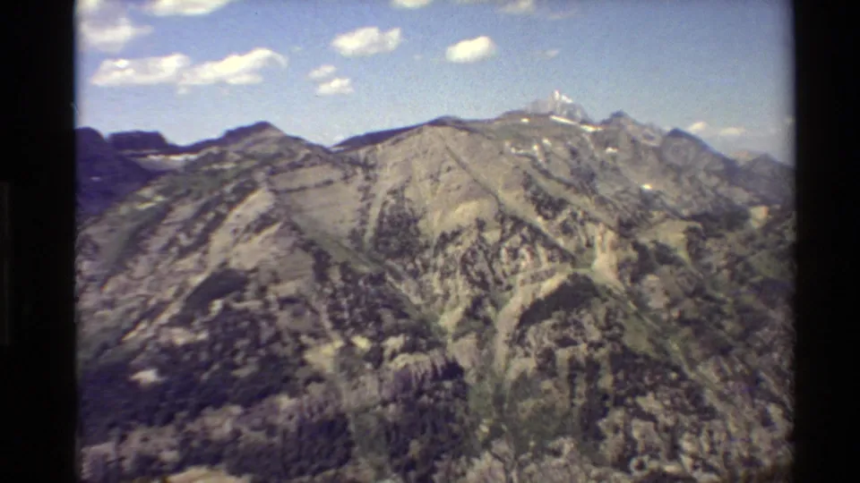 1980 Western USA home-movie frame ending on a rocky mountain shoulder with evergreen patches, distant snow, and small clouds overhead.