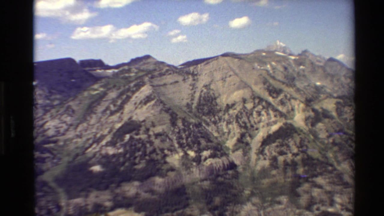 1980s 8mm Western USA mountain view showing rugged ridges, forested slopes, lingering snow patches, and scattered clouds.