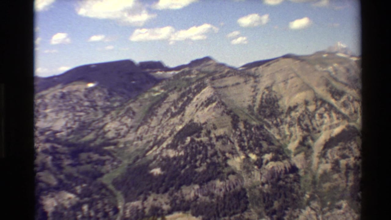 Western USA 8mm frame of a broad mountain range with rocky faces, evergreen patches, snow flecks, and a bright clouded sky.