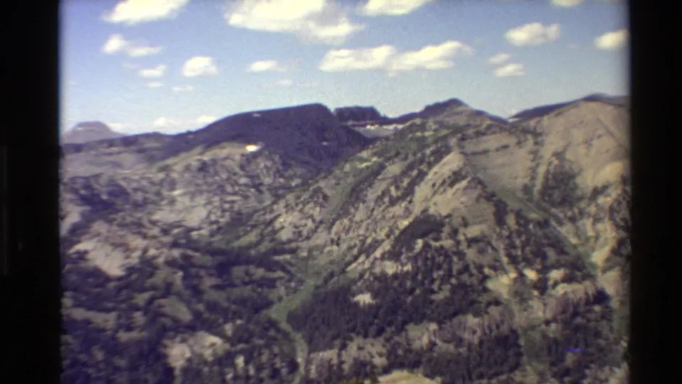 1980s home-movie view across Western USA mountains, with distant rounded peaks, rocky ridges, tree patches, and open sky.