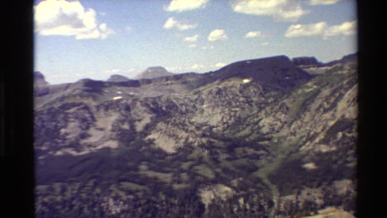 Wide mountain range under blue sky with scattered clouds and rocky forested slopes.