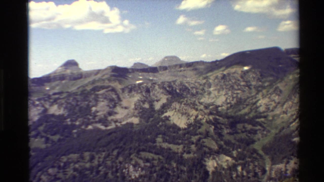 Western U.S. mountain panorama on 8mm film, with rocky ridgelines, green valleys, a rounded peak, and bright scattered clouds.