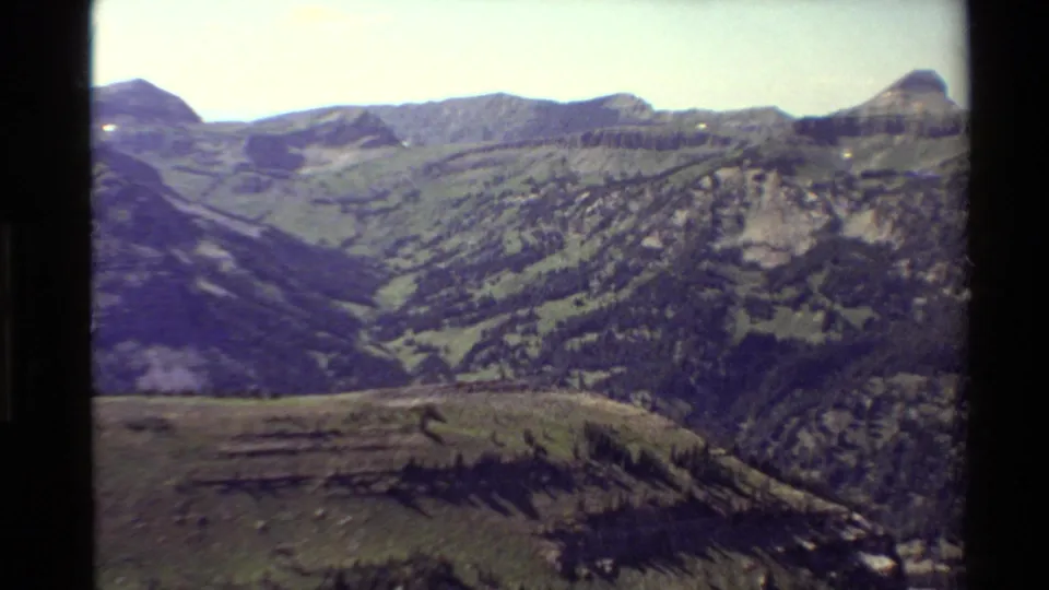 1980 8mm home-movie view across a foreground ridge toward layered mountain slopes, forested valley areas, and distant peaks.