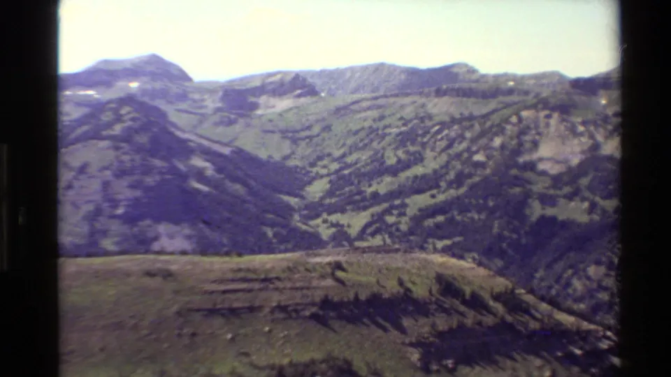 Western U.S. archival 8mm frame showing an open mountain valley, textured ridges, forested patches, and distant alpine peaks.