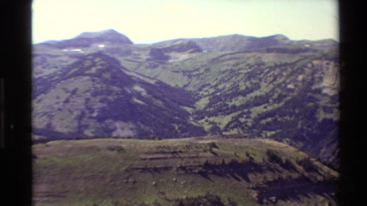 1980 Western USA 8mm frame showing layered mountain ridges, green slopes, and a bright sky from a high scenic viewpoint.