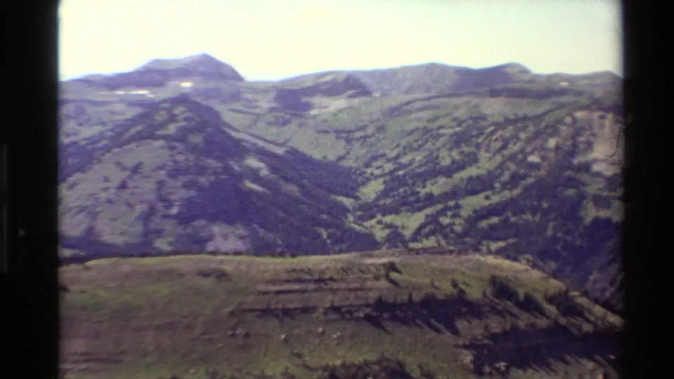 1980 Western USA 8mm frame showing layered mountain ridges, green slopes, and a bright sky from a high scenic viewpoint.