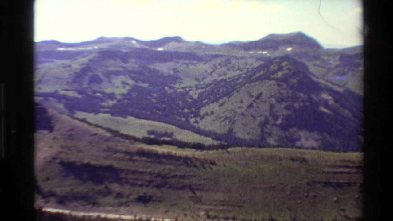 1980 Western USA 8mm frame showing grassy mountain ridges, scattered conifers, and a dirt road near the foreground.