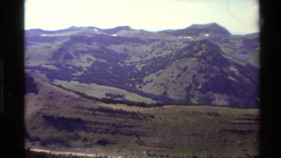 1980 Western USA 8mm frame showing grassy mountain ridges, scattered conifers, and a dirt road near the foreground.