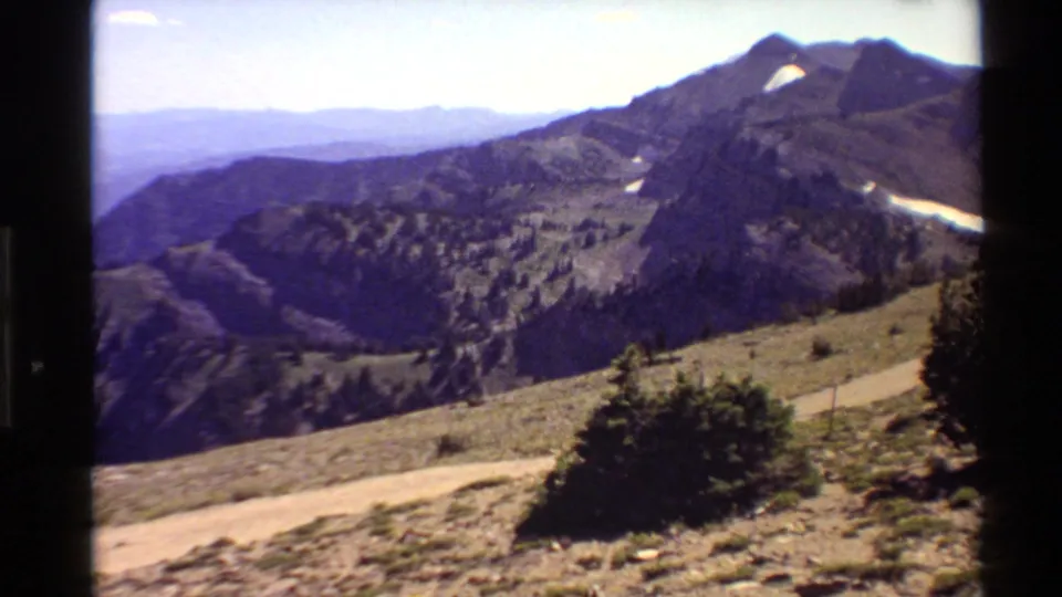 1980 Western USA 8mm frame of a steep mountain landscape with conifers, road, distant valley, and lingering snow patches.