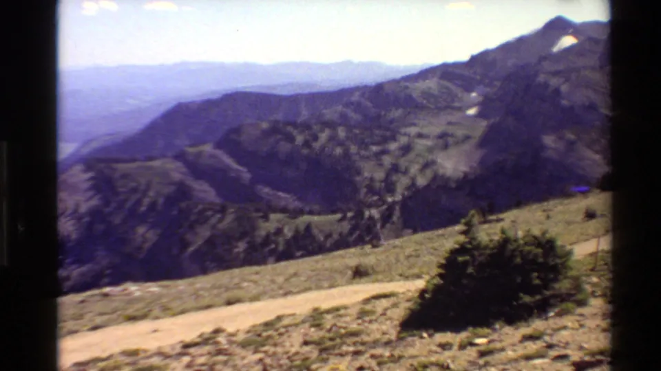 1980 Western USA archival frame showing a foreground road, conifered slopes, distant ridges, and small snow patches near the peaks.