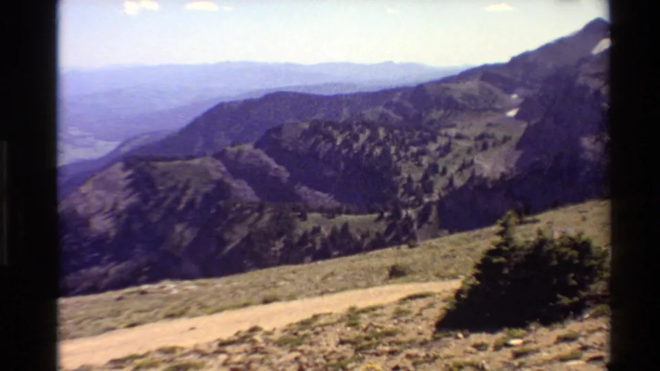 1980 Western USA 8mm frame showing rugged mountain ridges, a foreground dirt walkway, sparse alpine vegetation, and distant hazy terrain.